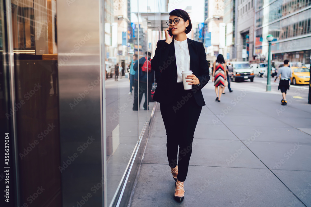 Pensive businesswoman in stylish formalwear strolling with coffee to go cup talking on phone about work, confident young female entrepreneur walking in downtown having serious mobile conversation. Pensive businesswoman in stylish formalwear strolling with coffee to go cup talking on phone about work, confident young female entrepreneur walking in downtown having serious mobile conversation.