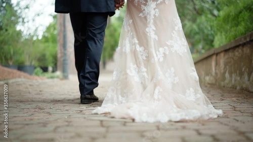 Close-up of fashion lace wedding dress of milk color stretching along the ancient cobblestone in the park. Just married couple walking slowly in the garden after the wedding ceremony holding tenderly