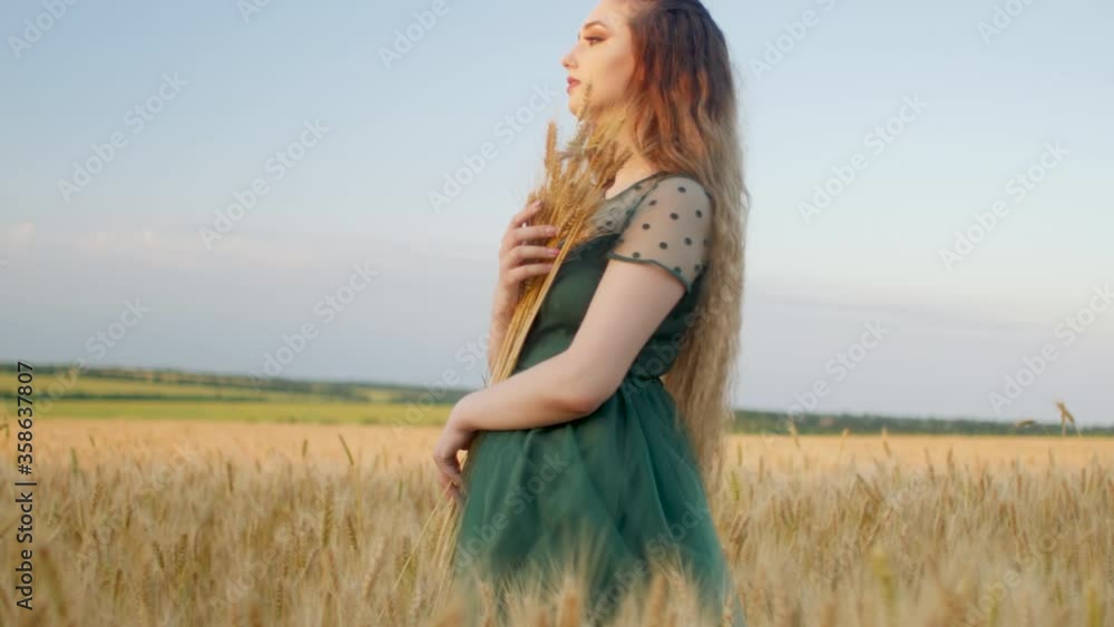 Beautiful woman in green dress on field with sheaf of wheat in hand at sunset light, girl enjoying summer nature landscape, harvest Beautiful woman in green dress on field with sheaf of wheat in hand at sunset light, girl enjoying summer nature landscape, harvest
