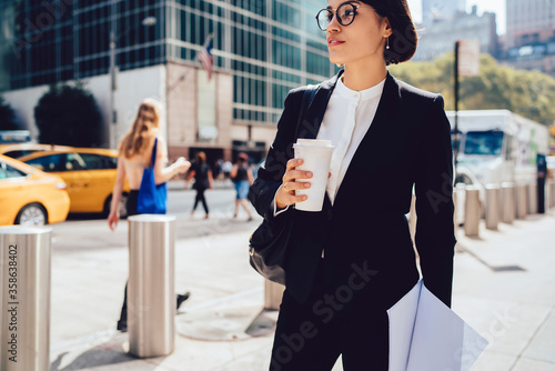 Foto Thoughtful female entrepreneur in trendy eyeglasses getting to work by foot in morning holding mock up coffee to go cup, pensive businesswoman in elegant formal wear strolling on street in downtown