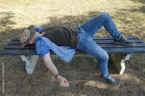 Man in jeans laying on bench in shadow with hat on his face. His arm hanging down with cigarette