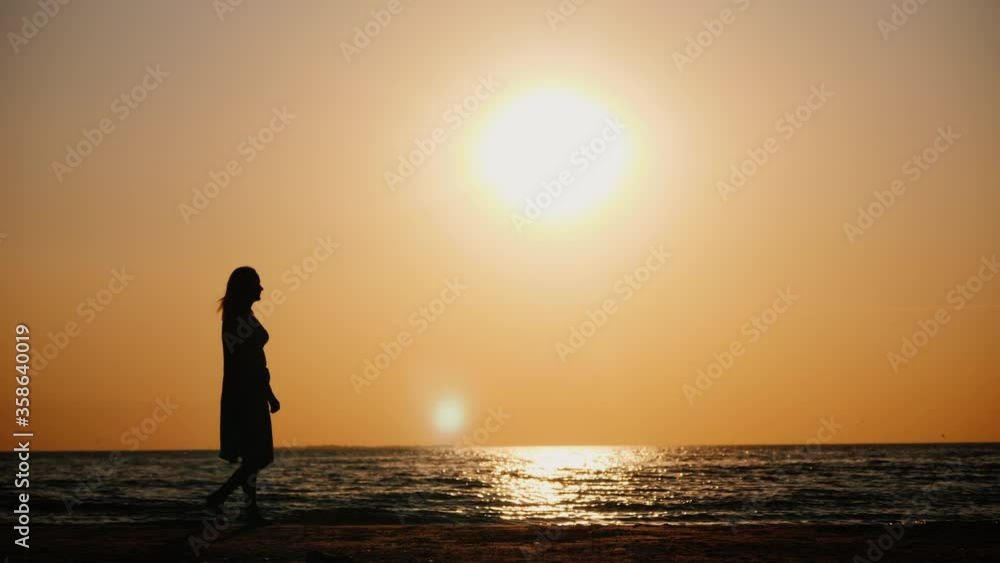 The silhouette of a woman who walks along the shore of the sea at sunset The silhouette of a woman who walks along the shore of the sea at sunset