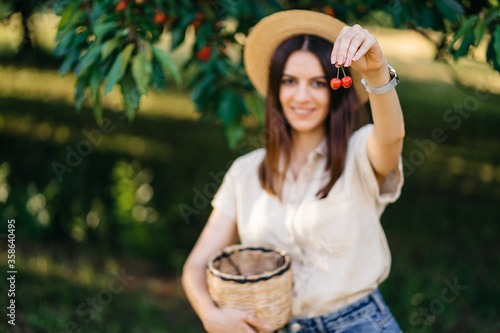 Portrait of a young cheerful woman in a straw hat with a basket  harvests sweet cherries in the garden. Agritourism Eco products