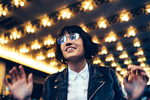 Young trendy dressed hipster in eyewear with night city light reflection fascinated with beautiful illumination, gorgeous woman in leather jacket wondering standing on urban background at evening