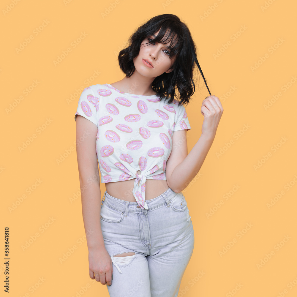 Fashionable brunette woman touching her hair and posing on a yellow studio wall in jeans and modern t-shirt Fashionable brunette woman touching her hair and posing on a yellow studio wall in jeans and modern t-shirt