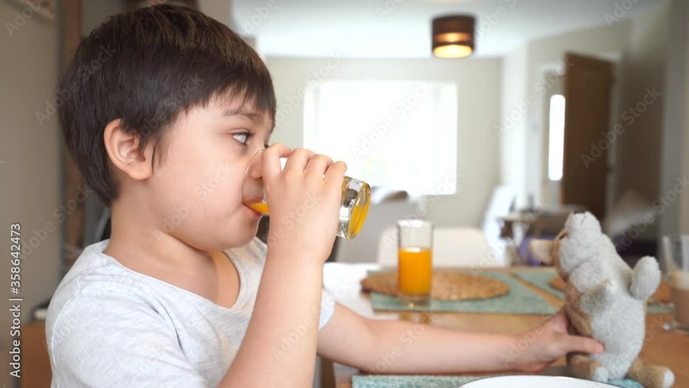 Healthy kid drinking fresh orange juice from clear glass and playing with his toy.New normal healthy lifestyle with child boy having breakfast with fesh food at home. Family time on dinning table Healthy kid drinking fresh orange juice from clear glass and playing with his toy.New normal healthy lifestyle with child boy having breakfast with fesh food at home. Family time on dinning table