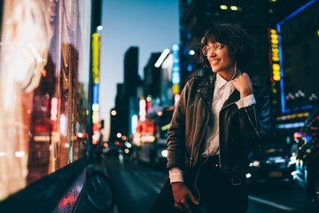 Smiling young woman in gorgeous apparel listening favorite song in earphones walking on night urban city street, happy hipster girl 20s enjoying music spending evening in illuminated Manhattan