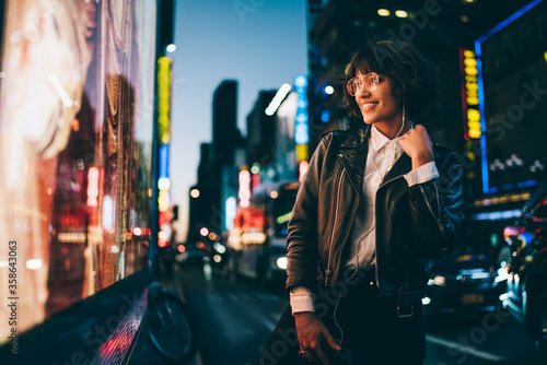 Smiling young woman in gorgeous apparel listening favorite song in earphones walking on night urban city street, happy hipster girl 20s enjoying music spending evening in illuminated Manhattan