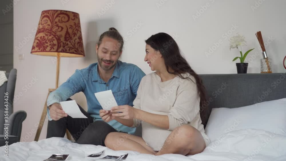 Beautiful Caucasian pregnant woman wife and her husband looking ultrasound baby picture together on the bed. Man touching his wife pregnant belly with love and smile. Happy family and new life concept Beautiful Caucasian pregnant woman wife and her husband looking ultrasound baby picture together on the bed. Man touching his wife pregnant belly with love and smile. Happy family and new life concept