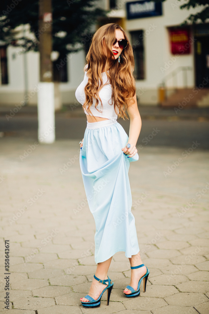 Stylish blonde girl in sunglasses with wavy hair tuned away and posing. Attractive woman smiling and holding her hair by hand. Wearing in top, mint skirt, make up with red lips. Walking in summertime.