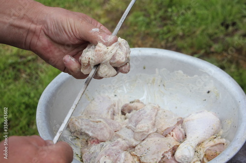 Men hand impales raw chicken meat on a spit from aluminium metal basin, outdoor cooking a poultru shashlik close up on green grass background on summer day
