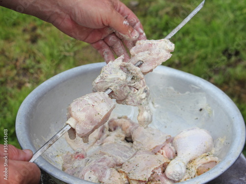 A rough male hand impales raw poultry meat on a skewer from alloy basin, outdoor cooking a chicken close up on green grass background on summer day photo