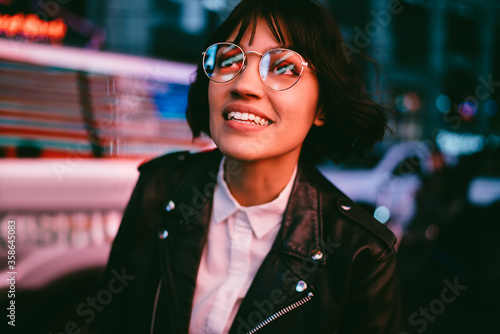 Konstfotografi Happy brunette young woman with short haircut looking up in stylish eyewear with night city light reflection fascinated with amazed illumination