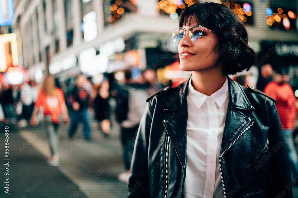 Fototapeta premium Carefree hipster tourist in optical spectacles for provide eyes protection enjoying evening walk in New York, attractive Caucasian woman dressed in trendy jacket looking around on Manhattan