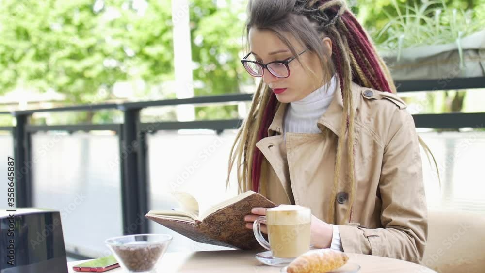 Modern young business woman with long dreadlocks and eyeglasses is writting some notes in her notebook while sitting in a street coffee shop during a day, remote work concept Modern young business woman with long dreadlocks and eyeglasses is writting some notes in her notebook while sitting in a street coffee shop during a day, remote work concept