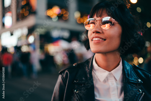 Carefree brunette young woman with short haircut in stylish eyewear for eyes protection fascinated with New York urbanity in Manhattan,youthful female in leather jacket enjoying metropolitan nightlife photo