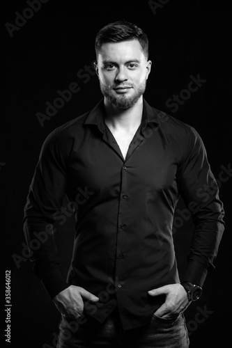 Young man in black shirt smiling isolated on dark background. Standing hands in pockets. Muscular male, athletic. Fashion portrait