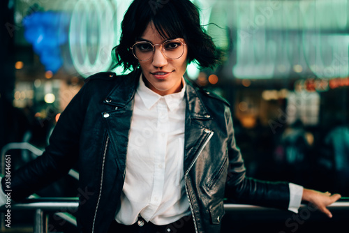 Half length portrait of attractive brunette hipster girl in eyeglasses dressed in casual stylish outfit looking at camera standing on blurred background in urban setting in evening leisure time