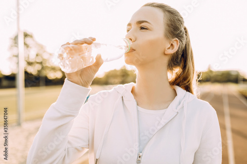 Athletic beautiful runner drinks water from a plastic bottle after running. A girl quenches her thirst after outdoor fitness classes photo