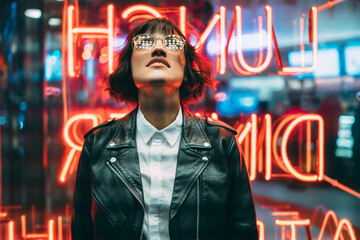 Stylish brunette woman in trendy apparel and eyewear looking up enjoying nightlife in city.Gorgeous hipster girl dressed in leather jacket standing outdoors on street with neon city illumination