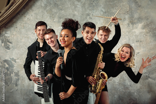 Cheerful international music group on a gray wall background, a group of musicians posing on camera in the hands of various instruments, guitars, saxophone. Copy space.
