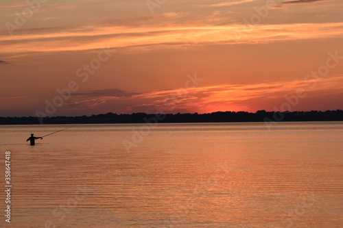 Fishing at sunset in the Eckernförde Bay, Northern Germany. The dark silhouette of a fisherman stands waist-deep in the Baltic Sea while the setting sun paints the sky and water in shades of orange