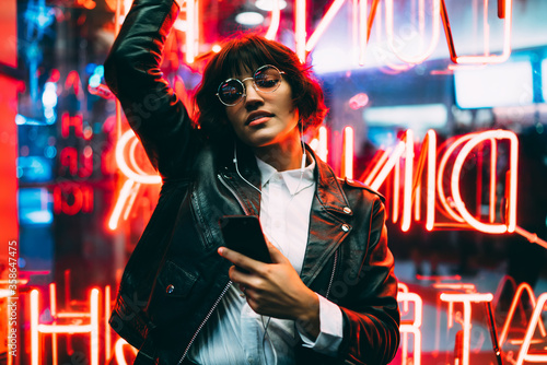 Beautiful brunette woman in trendy apparel and eyewear enjoying nightlife in city listening music in earphones and moving to sound, gorgeous hipster girl dancing outdoors on neon city illumination . photo