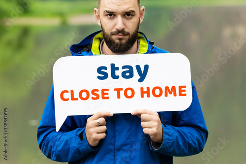 A young man in a blue jacket stands in the nature in summer and holds the message on the banner stay close to home. The concept of social distance in the fight against pandemic 2020. coronavirus photo