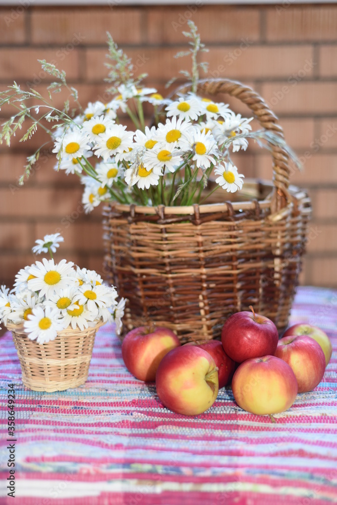 A large bouquet of daisies in a wicker basket, a small bouquet - in a pot on a woven striped bright carpet. Ripe juicy apples lie nearby. Vertical photo.