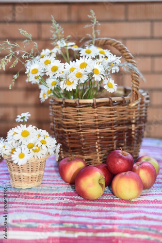 A large bouquet of daisies in a wicker basket  a small bouquet - in a pot on a woven striped bright carpet. Ripe juicy apples lie nearby. Vertical photo.