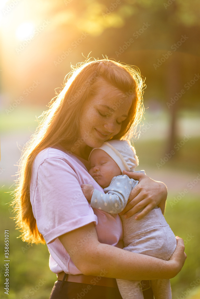 Young mother hugs a two-month-old newborn baby in the summer green park. Moments of happy motherhood - obrazy, fototapety, plakaty 
