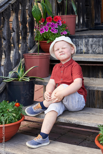 Little funny boy sits on the steps of an old house near potted flowers.