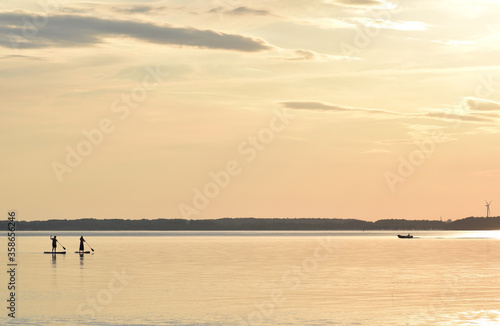 Paddling at sunset in the Eckernförde Bay, in Northern Germany. The dark silhouette of two people on paddling boards stand against the golden color of the Baltic sea.