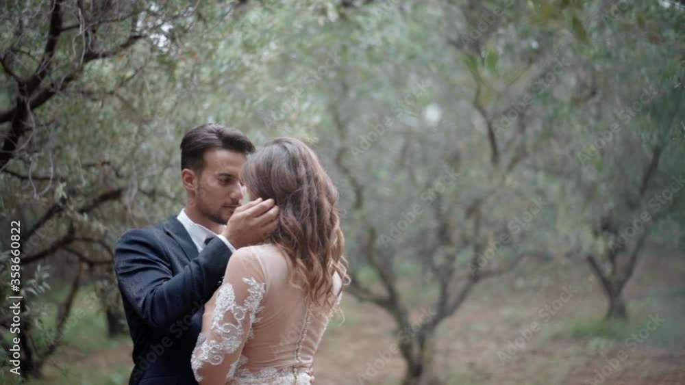 Amazing cinematic frame of young couple in love among the olive trees at the plantation. Handsome man in dark blue suit looking at his pretty lady in lace dress and touching her curly hair and Amazing cinematic frame of young couple in love among the olive trees at the plantation. Handsome man in dark blue suit looking at his pretty lady in lace dress and touching her curly hair and