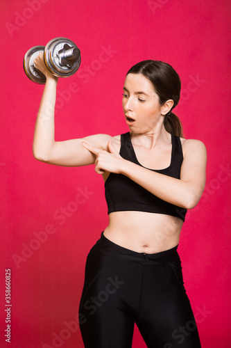 Beautiful woman doing exercises with dumbbells, pumping biceps. Photo of athletic woman in fashionable black sportswear on red background. Strength and motivation