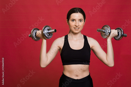 Sporty woman jumping with dumbbells. Photo of active woman in sportswear on red background. Dynamic movement. Sport and healthy lifestyle