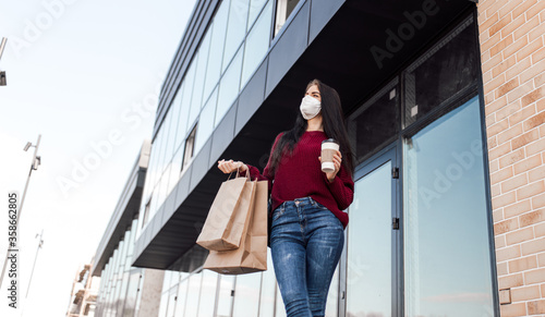 Young woman in medical mask walk outdoors with coffee cup and shopping bags after shopping