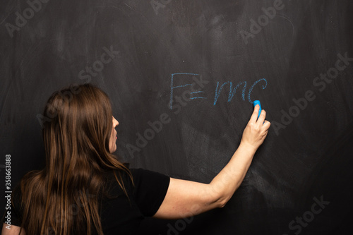 The physical formula of Einstein s theory is written by a young girl in blue chalk on a blackboard. FMC2 is written by a science teacher or student in the classroom.