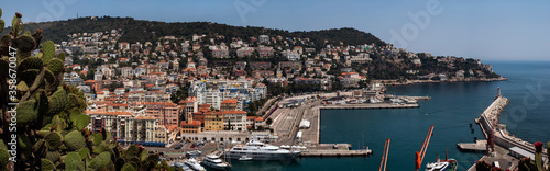 Fototapeta Naklejka Na Ścianę i Meble -  Panoramic View of Part of Nice City Port in France with Boats Berth Added Lighthouse Cape and City Buildings on Hill - Houses Yachts and Sea in Port Lympia in Summer