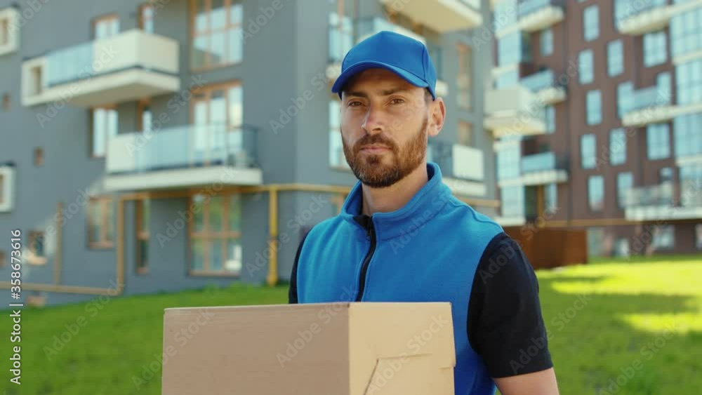 Serious handsome young delivery man holding cardboard box stand look at camera at street outdoor shopping package postman slow motion Serious handsome young delivery man holding cardboard box stand look at camera at street outdoor shopping package postman slow motion
