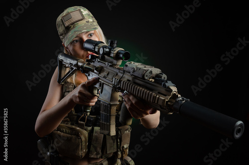 Fotografia a sexy girl in military airsoft overalls poses with a gun in her hands on a dark