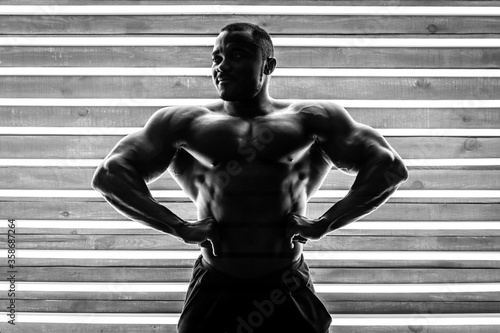 African male athlete demonstrates muscles on a background of wood and fluorescent lamps. Black and white photo