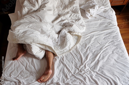 Close-up man feet alone in white bed, view of feet man sleeping and lying under white blanket and white bed , couples feet crossed under the duvet in bed