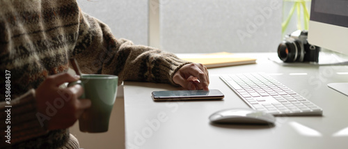Female employee relaxing with smartphone and holding a cup of coffee at office desk