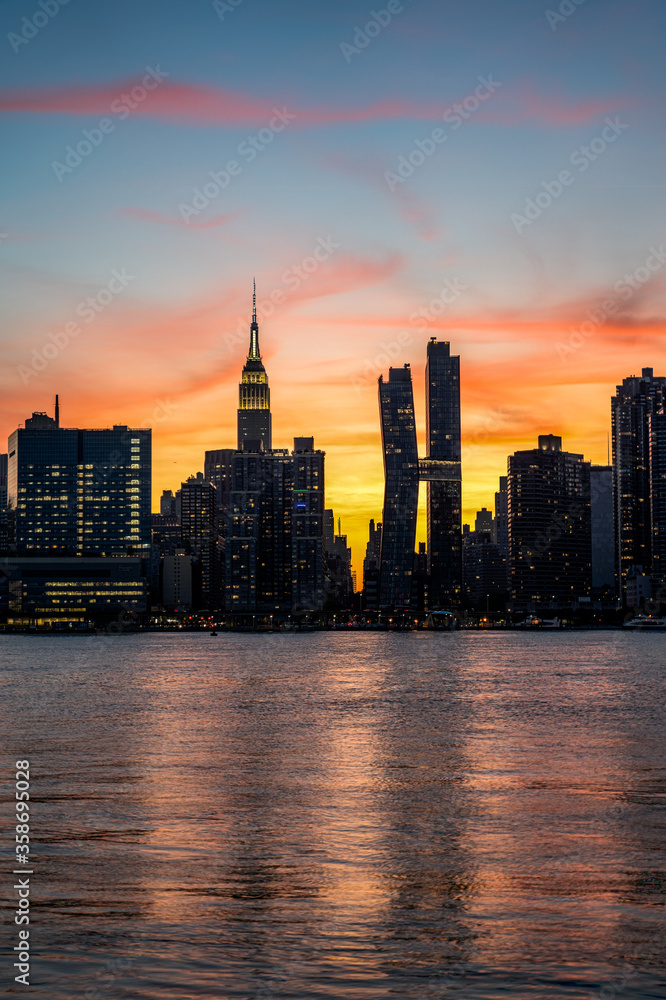 Manhattan Midtown skyline view. Dark silhouettes of Empire State building, and American Copper Buildings. Sunset, orange sky, reflections in water
