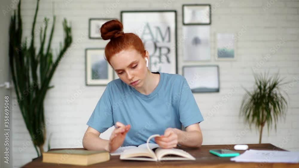 Serious redhead young woman student is flipping book looking for right page at home. Beautiful female wearing earphone is listening an audio lesson at desk in cozy living room in self-isolation. Serious redhead young woman student is flipping book looking for right page at home. Beautiful female wearing earphone is listening an audio lesson at desk in cozy living room in self-isolation.