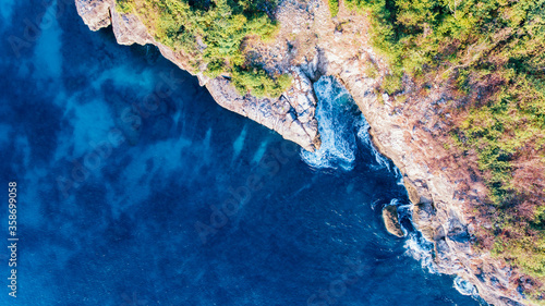 Aerial view of rocky hill coast with green trees and blue sea with white foaming waves in Sumbawa, Indonesia
