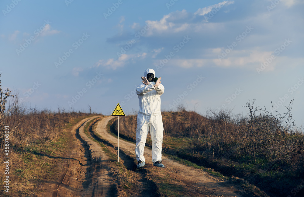 Obraz premium Scientist in radiation suit and gas mask showing that entrance on territory is forbidden, standing near yellow triangle with skull and crossbones sign and warning about poisonous substances and danger