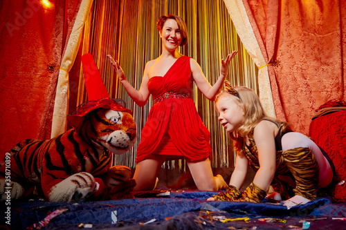 Family during a stylized theatrical circus photoshoot in a beautiful red location. Models mother who looks like a trainer and daughter who looks like a tiger pose on stage with curtains