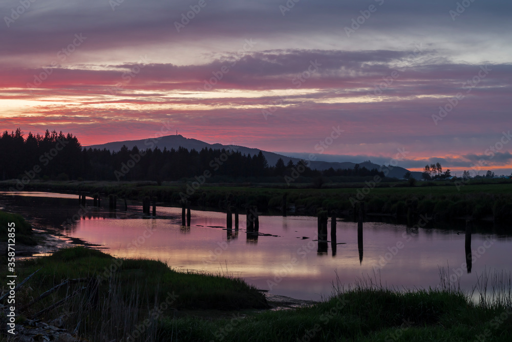 View of vibrant colors sunset landscape. Silhouetted hills and plants reflect in calm water of the river. Location is Tillamook river in Oregon, USA
